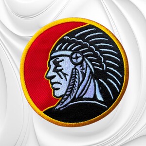 May include: A circular embroidered patch featuring a profile view of a person wearing a traditional feathered headdress. The design uses a color palette of black, white, and light blue against a vibrant red background, all enclosed by a yellow border.
