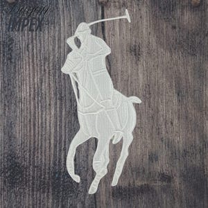 May include: White embroidered patch featuring a polo player on a horse, holding a mallet. The patch is a detailed outline of the iconic logo. The patch is on a wooden background.