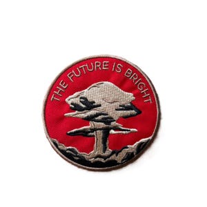 May include: A circular embroidered patch featuring a red background with a beige and black mushroom cloud illustration in the center. The text THE FUTURE IS BRIGHT is stitched in beige along the upper curve of the patch, which has a beige border.