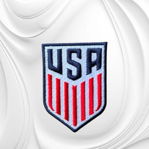 May include: A shield-shaped embroidered patch featuring the letters USA in dark navy blue at the top, positioned above a series of vertical red stripes on a white background, all enclosed within a dark navy blue border with a light blue outline.