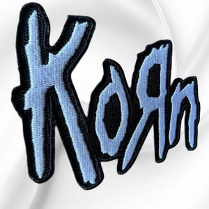 May include: An embroidered patch featuring the band name Korn in a stylized, jagged, light blue font with a thick black border. The lettering has a distressed, rough-edged appearance, and the entire design is set against a solid black background.