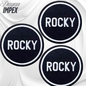 May include: Three black circular patches with the word "ROCKY" in white block letters. Each patch has a white border. The patches are arranged on a white and gray background. The text "Dayan IMPEX" is in the upper left corner.