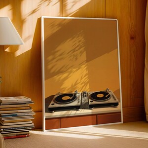 A white-framed poster of DJ turntables stands on the floor against a wood-paneled wall next to a stack of books and a lamp.