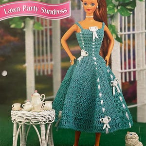 Pode incluir: Uma boneca vestindo um vestido de verão de crochê turquesa com detalhes de fita branca e laços. A boneca está posando em frente a uma treliça branca. O texto "Lawn Party Sundress" é visível. Uma pequena mesa branca com um jogo de chá e uma estatueta de cachorro também estão presentes.