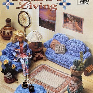 May include: A miniature crocheted living room set, including a blue sofa, rug, and table, with a doll seated on the sofa. The scene is decorated with small artworks and a lamp. The words "Santa Fe Living" are displayed at the top of the image.