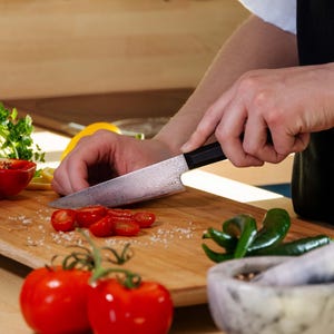 Peut inclure: Une personne coupe des tomates sur une planche à découper en bois avec un couteau à motifs. La scène comprend des tomates fraîches, des poivrons verts et un mortier et un pilon, suggérant la préparation des aliments dans une cuisine.