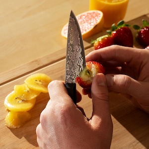 Peut inclure: Un couteau en acier inoxydable à lame à motifs est utilisé pour couper une fraise. Des kiwis tranchés et un verre de jus d'orange sont également sur la planche à découper en bois. Des fraises fraîches et une moitié de pamplemousse sont également visibles.