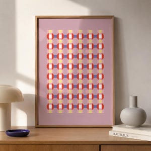 Puede incluir: Una lámina de arte enmarcada con un patrón geométrico repetitivo de cuadrados y octágonos rojos, azules, amarillos y blancos sobre un fondo malva sólido. La obra está contenida en un marco de madera de tono claro con un perfil fino.
