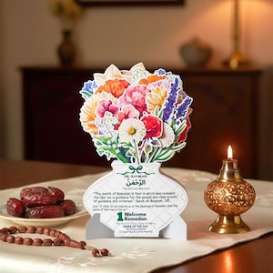 Könnte beinhalten: Eine dekorative Ramadan-Grußkarte mit einem bunten Blumenarrangement in Vasenform. Die Karte enthält arabischen und englischen Text, eine brennende Kerze, Datteln und Gebetsketten. Die Karte liegt auf einem Tisch.