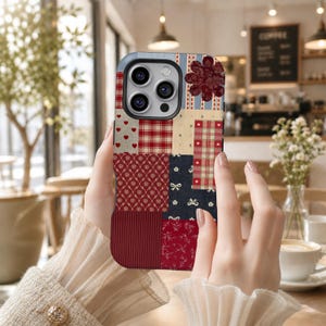 Puede incluir: Una funda de teléfono patchwork con una variedad de patrones que incluyen corazones, cuadros y diseños florales en rojo, crema y azul marino. La funda tiene un acento de flor rojo oscuro y una sección inferior texturizada y acanalada. La funda del teléfono es sostenida por una persona.