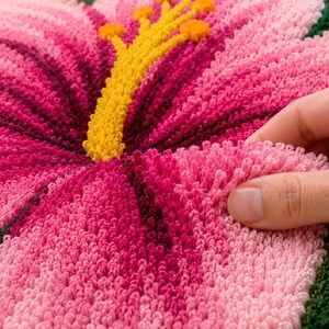 May include: Close-up of a handmade, pink hibiscus flower rug. The rug features a detailed design with varying shades of pink, a yellow centre, and green leaves. The texture is soft and plush, with a hand touching the petals.
