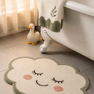 May include: A cloud-shaped bath mat in cream with a sage green border, featuring a smiling face with closed eyes and pink cheeks. A matching cream towel with a sage green scalloped edge and a small embroidered leaf sprig hangs nearby.