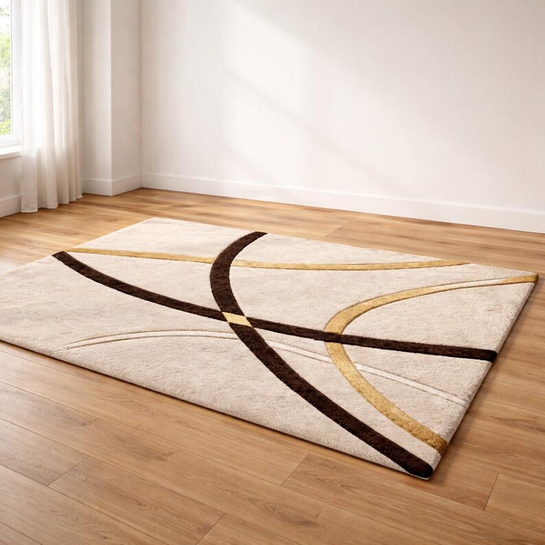 May include: Beige rectangular area rug with a modern design. The rug features intersecting curved lines in brown and gold against a neutral background. The rug is placed on a hardwood floor.
