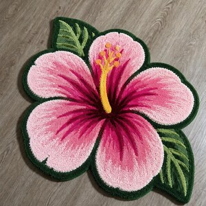 May include: A pink and magenta hibiscus flower rug with green leaves and a dark green border. The rug is placed on a wooden floor. The flower's centre is yellow and the petals have a gradient of pink and magenta.