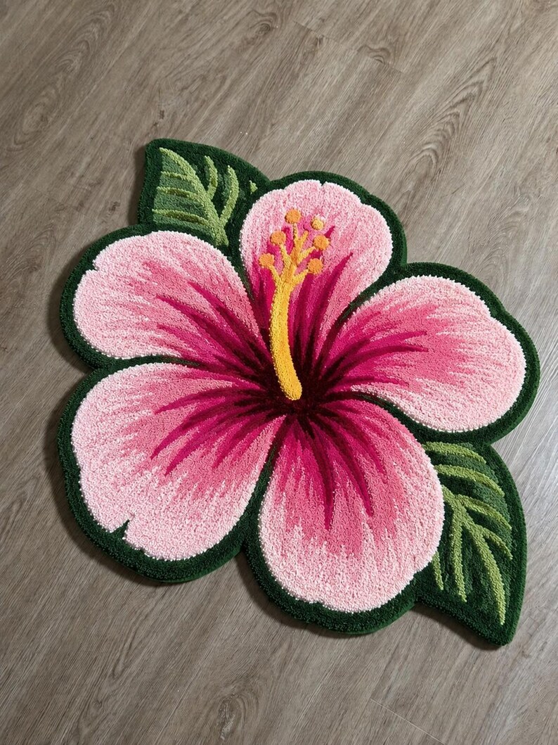 May include: A pink and magenta hibiscus flower rug with green leaves and a dark green border. The rug is placed on a wooden floor. The flower's centre is yellow and the petals have a gradient of pink and magenta.