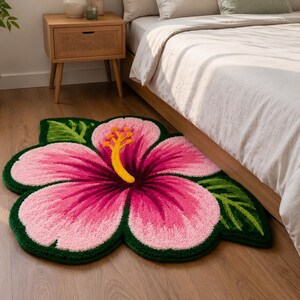 May include: A colourful, flower-shaped rug with a pink hibiscus design, yellow stamen, and green leaves. The rug is on a wooden floor near a bed, adding a tropical touch to the room's decor. The rug's dimensions are approximately 120 cm.