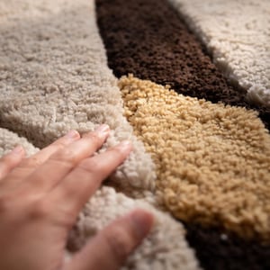 May include: Close-up of a textured rug with a geometric pattern. The rug features a combination of beige, brown, and tan colors. The soft, plush texture is visible, with a hand gently touching the surface.