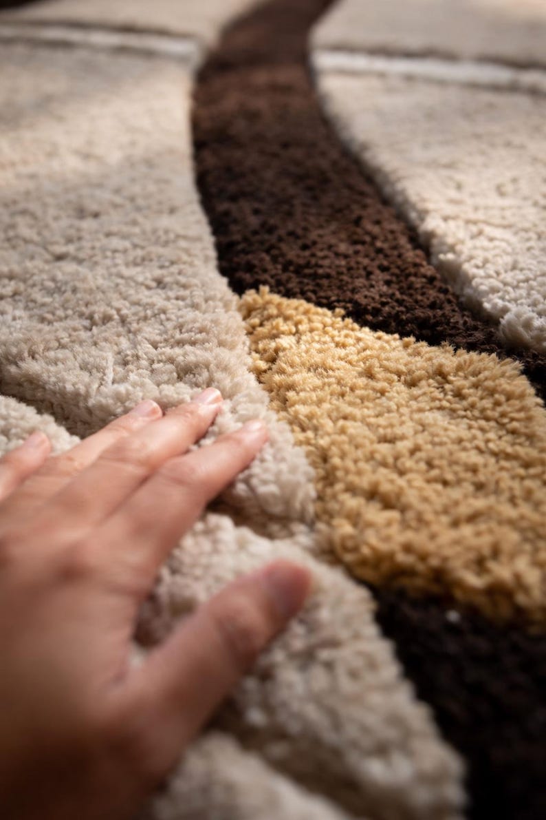 May include: Close-up of a textured rug with a geometric pattern. The rug features a combination of beige, brown, and tan colors. The soft, plush texture is visible, with a hand gently touching the surface.