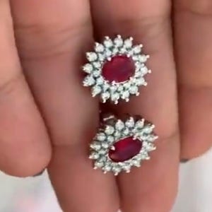 Peut inclure: Une paire de boucles d'oreilles à clous avec des pierres précieuses ovales rouge foncé, entourées de petites pierres précieuses claires. Les boucles d'oreilles ont un design floral et sont serties dans un métal argenté. Accessoire de bijouterie.