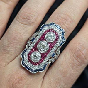 May include: An Art Deco ring with three large, round diamonds in a rectangular setting. The ring features a central panel of red stones, framed by blue and white accents, and detailed metalwork.