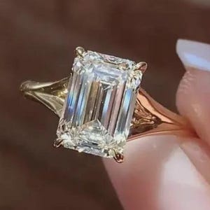 Könnte beinhalten: Ein goldener Ring mit einem großen, rechteckig geschliffenen Diamanten. Der klare Diamant reflektiert das Licht und ist in einer goldenen Fassung mit geteiltem Schaftdesign gefasst. Der Ring wird vor einem verschwommenen Hintergrund gehalten.