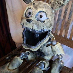 May include: A detailed, articulated, and weathered rabbit-like animatronic figure with a wide open mouth revealing sharp teeth. The figure is primarily cream and grey, with visible mechanical joints and a distressed appearance. It sits on a wooden chair.