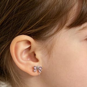 以下が含まれることがあります： 子供の耳のクローズアップ。銀色のリボン型のイヤリングが付いています。イヤリングはピンクのエナメル充填と中央に小さな透明な宝石が付いています。イヤリングはクリップオンタイプです。