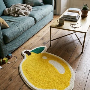 May include: A yellow lemon-shaped rug with a white border and green leaf detail rests on a hardwood floor. The rug is in a living room setting, near a teal sofa and a gold coffee table.