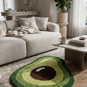 May include: Avocado-shaped rug with varying shades of green and a brown pit. The rug is placed on a wooden floor in a living room setting, near a beige sofa and a marble coffee table.