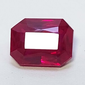 May include: A vibrant, deep red gemstone cut into a rectangular emerald shape. The stone features multiple facets that reflect light, creating a brilliant, translucent appearance with a clear, bright white reflection visible on its top surface.