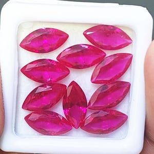 Pode incluir: Uma coleção de nove pedras preciosas rosa vibrantes, cortadas em forma de marquesa, dispostas em um recipiente de plástico branco. As pedras são facetadas, refletindo a luz. O recipiente é quadrado.