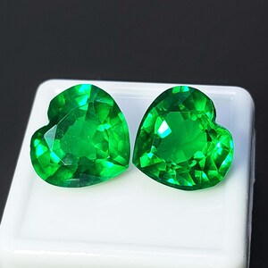 May include: Two heart-shaped, faceted gemstones in a vibrant green hue. The gems are displayed on a white surface against a dark background. The cut of the stones creates multiple reflective surfaces.