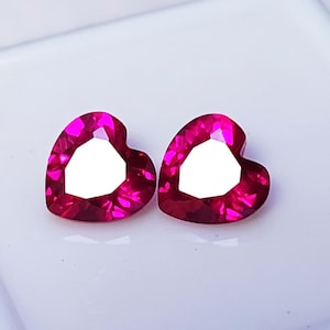 May include: Two heart-shaped, faceted gemstones in a vibrant magenta hue. The gems have a bright, white center and are set against a white background. These gemstones could be used for jewelry making.