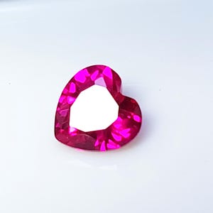 May include: A faceted, heart-shaped gemstone in a vibrant magenta hue. The gem has a bright, reflective surface with multiple facets, creating a sparkling effect. The heart shape is clearly defined, with a large, open space in the center.