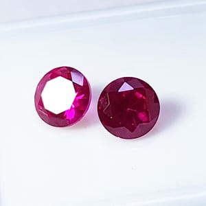 May include: Two round, faceted rubies with a deep red hue are displayed against a white background. The gemstones have a brilliant cut, showcasing their internal structure and light reflection. The rubies are a vibrant, saturated color.