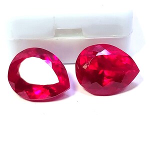 May include: Two faceted, pear-shaped rubies. One ruby has a hollow center, while the other is solid. Both gems are a deep, vibrant red color and are displayed against a white background.