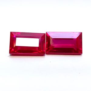 May include: Two rectangular, faceted, bright red gemstones. One gemstone has a rectangular hole in the center. The gems are set against a white background. The gems are likely rubies or similar precious stones.