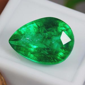 Peut inclure: Une émeraude en forme de poire, d'un vert vif. La surface à facettes reflète la lumière, révélant des inclusions internes. La gemme est posée sur un fond blanc, mettant en valeur son éclat. Vue détaillée des facettes.