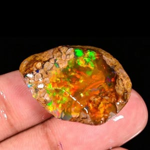 Puede incluir: Una vibrante gema de ópalo en bruto con un exterior rugoso y texturizado. El ópalo muestra un deslumbrante juego de colores, con destellos de verde, naranja y rojo. La gema se sostiene en una mano sobre un fondo oscuro.