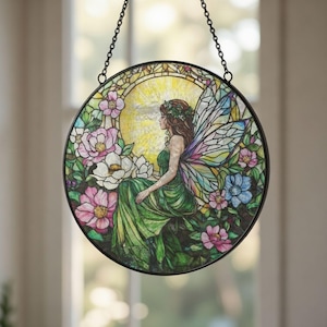 May include: Round stained glass window art featuring a fairy in a green dress with floral wings, surrounded by pink, white, and blue flowers. The fairy is seated in a garden setting with a yellow sun in the background. The window is framed with a black metal border.