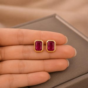 May include: A pair of stud earrings featuring rectangular, emerald-cut deep red gemstones set in polished yellow gold bezels. The stones have a vibrant, saturated crimson hue, and the metal frames provide a smooth, clean border around each gem.