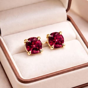 Pode incluir: Um par de brincos de ouro com grandes pedras preciosas redondas e vermelho escuro. Os brincos são colocados em uma configuração de ouro de quatro pontas e são exibidos em uma caixa de joias branca com uma borda marrom.
