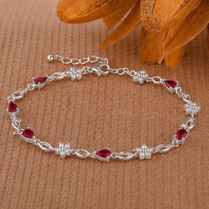 May include: A silver-toned bracelet featuring alternating teardrop-shaped red stones and small flower-shaped clusters of clear white stones. The chain is composed of delicate metal links and includes an adjustable extender with a small round bead.
