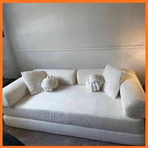 May include: A white corduroy sofa with matching pillows and knot-shaped decorative cushions. The sofa has a low profile and a minimalist design, perfect for a modern living space. The neutral color palette creates a calming and inviting atmosphere.