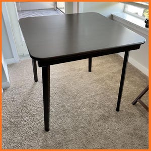 May include: A dark brown square table with four legs. The table has a smooth surface and rounded corners. The legs are also dark brown and appear to be made of wood. The table is approximately 30 inches square.