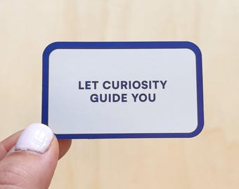 Op de afbeelding: Een rechthoekige sticker met afgeronde hoeken, met een witte achtergrond en een marineblauwe rand. De tekst "LET CURIOSITY GUIDE YOU" is gecentreerd in marineblauw. De sticker wordt vastgehouden door een hand met gelakte nagels.
