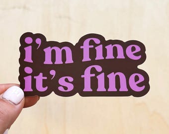 Op de afbeelding: Een sticker met de tekst "i'm fine it's fine" in lavendelkleurige tekst op een donkerbruine achtergrond. De sticker wordt vastgehouden door een hand met lichtroze nagellak.