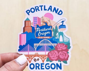 Op de afbeelding: Een kleurrijke sticker met bezienswaardigheden van Portland, Oregon. Het ontwerp bevat een brug, gebouwen, een berg en een kabelbaan, met de woorden "Portland Oregon" en "Old Town". Roze rozen staan onderaan.