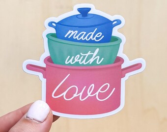 Op de afbeelding: Een sticker met drie gestapelde kookpotten in blauw, groen en roze. De woorden "made with love" staan in een sierlijk lettertype over de potten geschreven. De sticker wordt door een hand tegen een lichte houtnerf achtergrond gehouden.