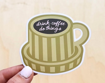 Op de afbeelding: Een witte keramische mok met een zwarte handgreep en de tekst "But First, Coffee" in een vetgedrukte, zwarte letter. De mok staat tegen een vage achtergrond. De mok is ongeveer 10 cm hoog.